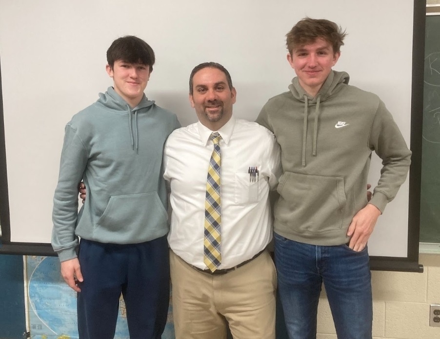 two senior boys in green sweatshirts standing on either side or Mr. Corbett
