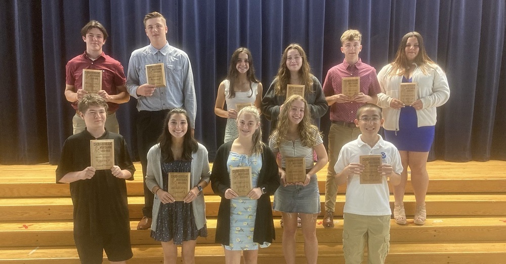male and female high school students on auditorium risers and holding sports awards plaques