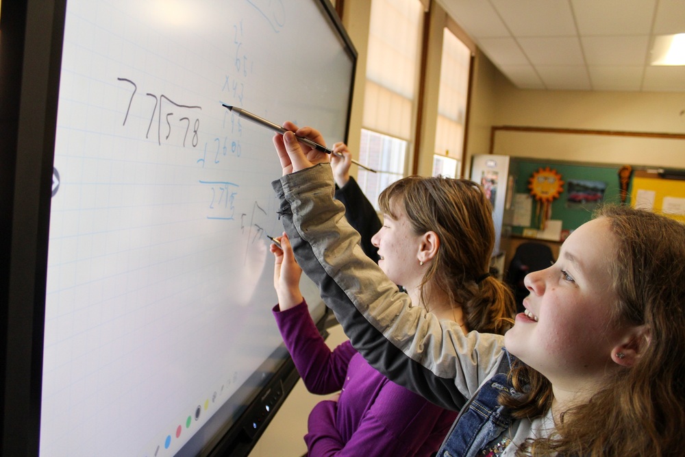 two female elementary students work on a long division problem using an interactive whiteboard