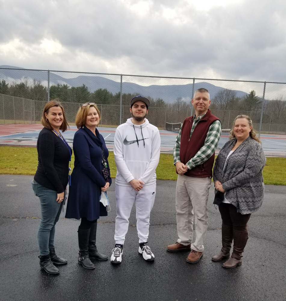 high school student dressed in white sweats with two teachers standing on either side of him while outdoors