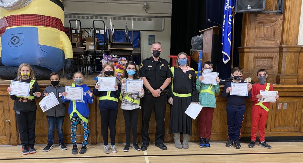 Deputy Espel and teacher stand in front of the gym stage with eight students wearing green safety belts and holding certificates