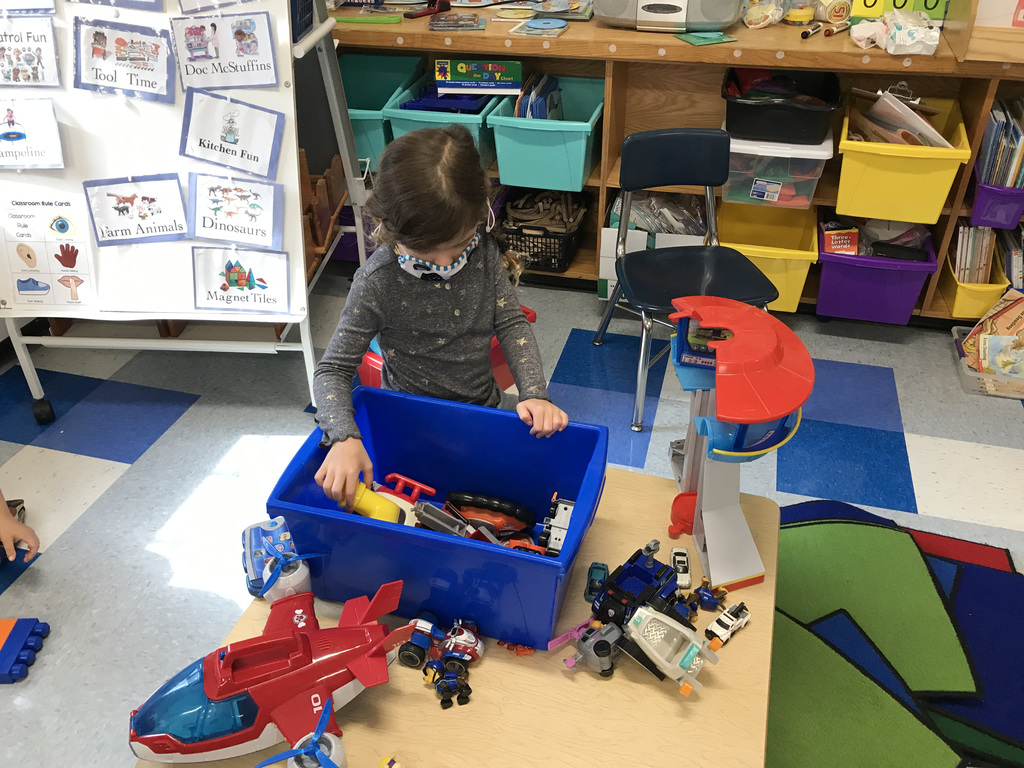 pre-kindergarten girl kneels on the classroom floor and removes toys from a blue bin
