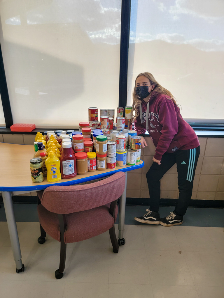 a high school girl stands next to a table that has non-perishable food items stacked on it
