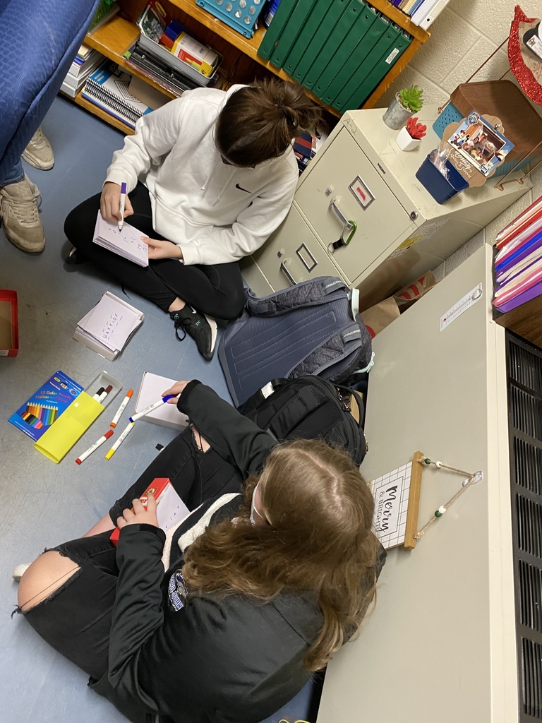 two students seated on a classroom floor writing and decorating cards