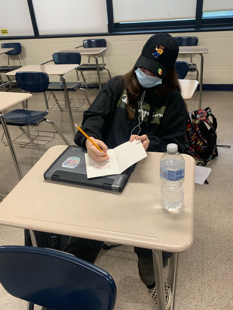 a high school student seated at a classroom desk writing a card