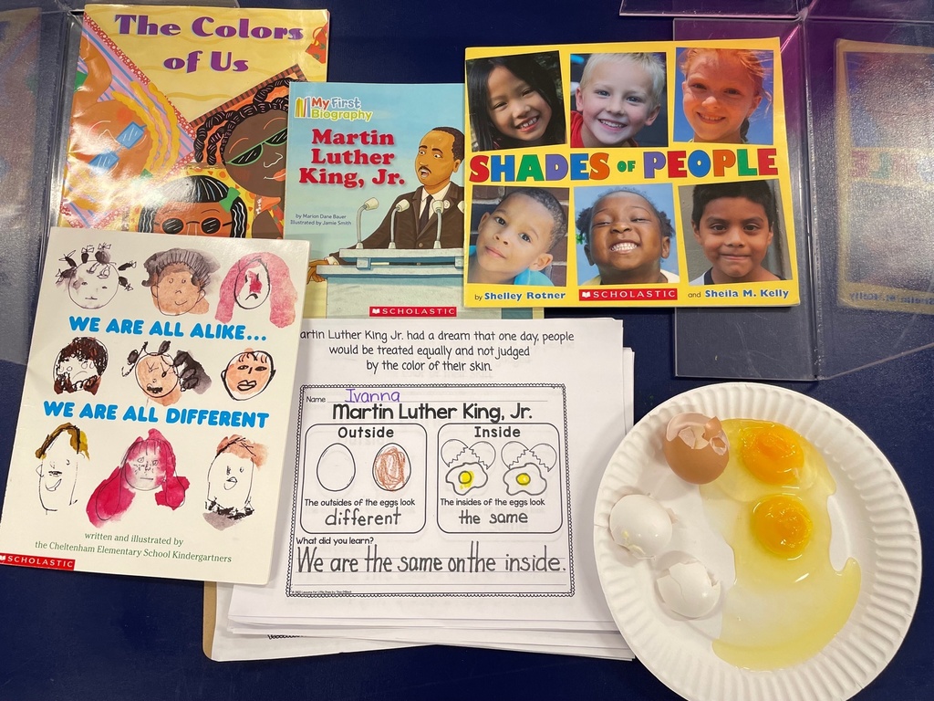 items on a desk depicting children with different hair and skin tones, and a paper plate with two cracked eggs one with a white shell and one with a brown shell