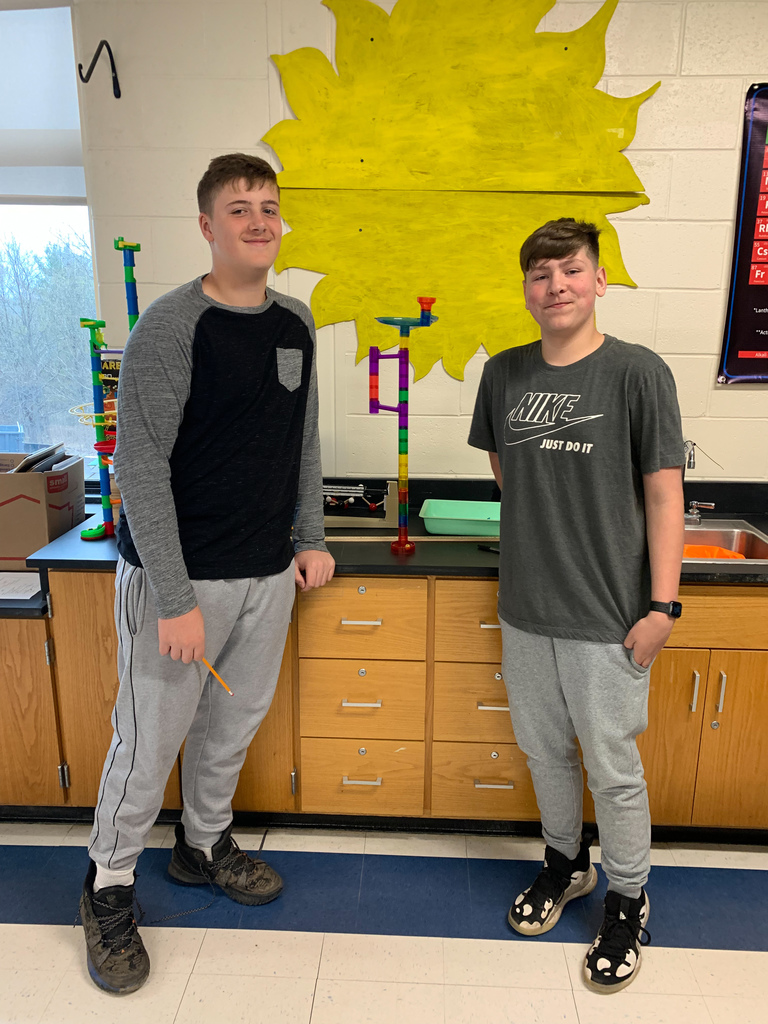 two middle school boys standing next to a counter that holds a plastic "marble run" to demonstrate potential energy; a yellow sun made of construction paper is on the wall behind them