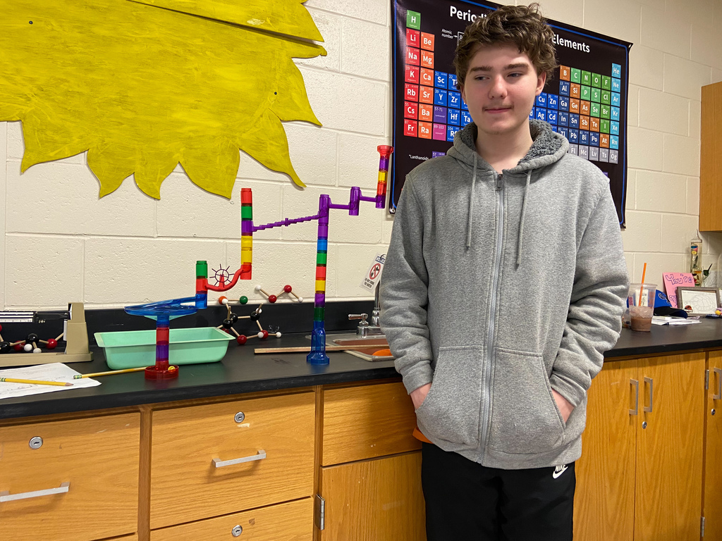 a boy in a gray sweatshirt stands next to a counter that holds a plastic "marble run" to demonstrate potential energy; a poster of the periodic table of elements is on the wall behind him