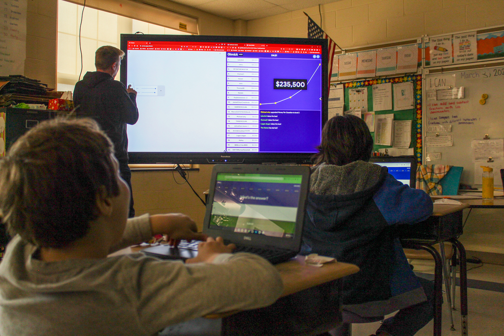 the back of the heads of two boys seated at classrooms desks using Chromebooks and interacting with content on an interactive white board that a teacher touches with his hand