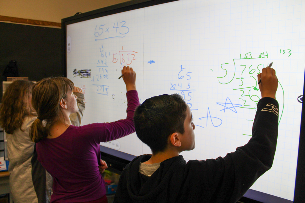 two girls and a boy work on long division problems using an interactive whiteboard