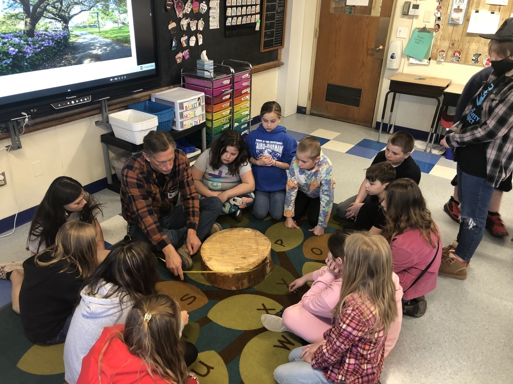 a group of elementary students sit in a circle on a classroom rug while an older man shows them a piece of a tree trunk and measures it