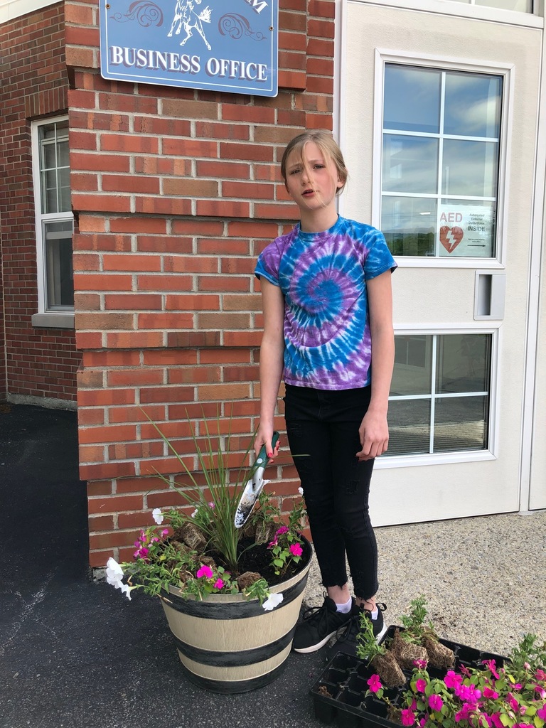 an elementary school girl in a blue and purple tie dyed t-shirt holding a hand shovel next to a wooden planter with flowers