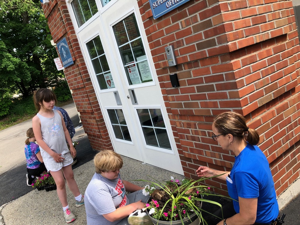 an elementary school boy helps a female custodian add flowers to a wooden planter in front of the school while an elementary school girl stands and watches