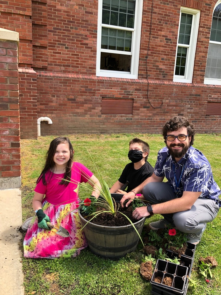 an elementary school girl and boy help Mr. Heilmann add flowers to a wooden planter in front of the school