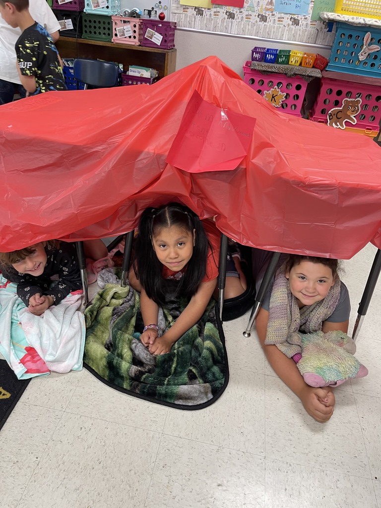 three first grade girls under their desks that are draped with red fabric to resemble a tent