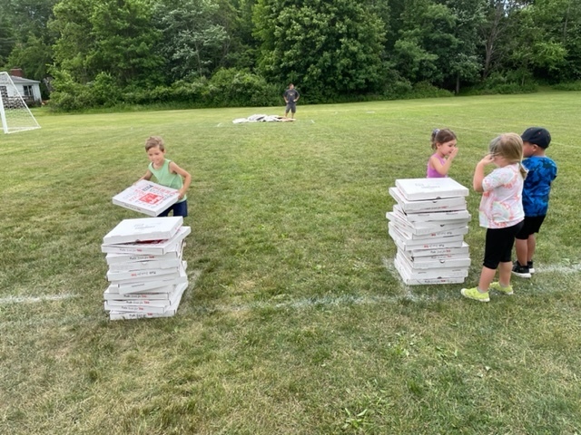 a pre-kindergarten boy stacks empty pizza boxes outside during field day