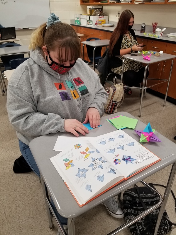 a high school girl seated at a classroom desk making complex origami from multicolored paper as she views an origami book